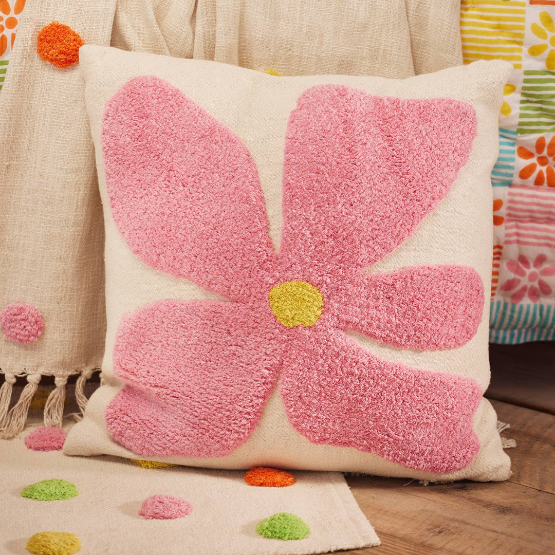 Decorative cushion with a bold pink flower design on a natural cotton cover, displayed against a bed with colorful bedding.