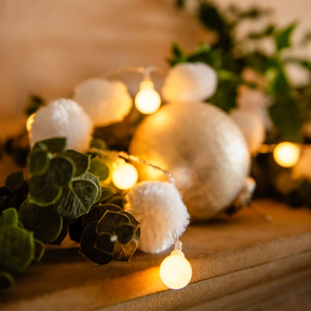 Decorative setup with white pom-pom string lights and Christmas greeneryon a wooden surface.