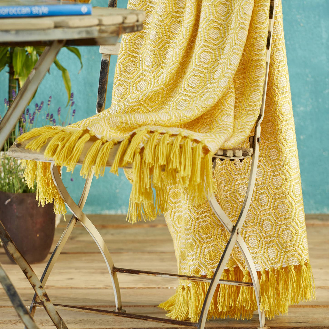A bright yellow woven throw blanket with a geometric pattern and long fringe, draped over a folding chair in an outdoor patio setting.