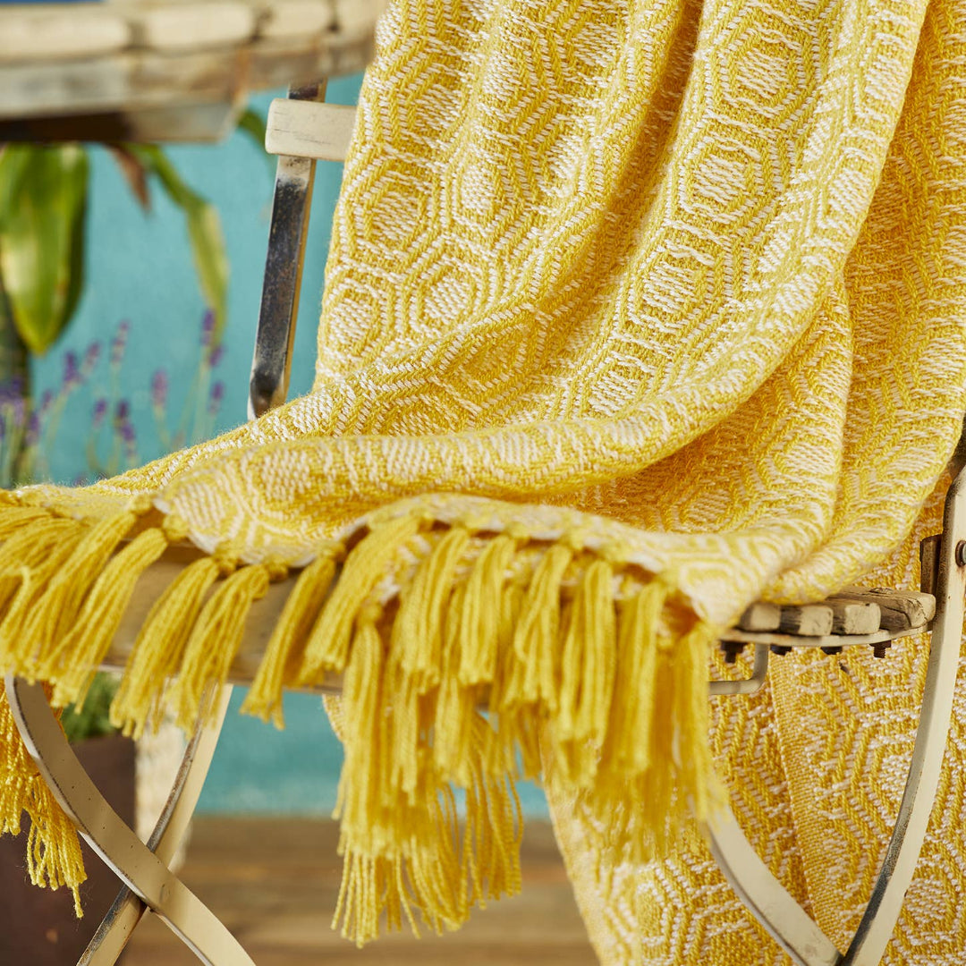 A bright yellow textured throw blanket with fringe tassels draped over a white wooden chair in a styled indoor setting.