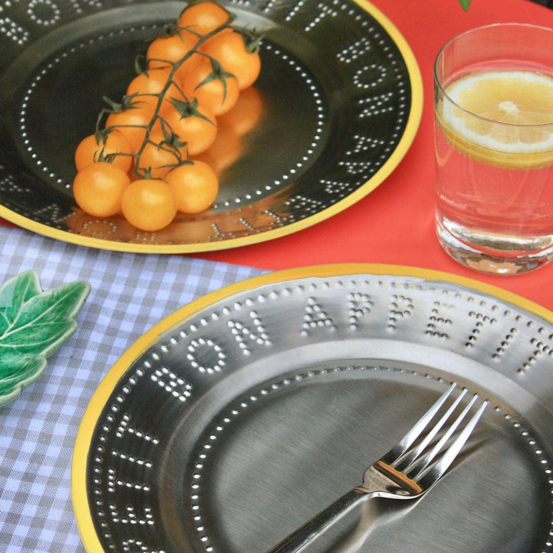 Two decorative steel plates with yellow trim embossed lettering and dotted borders, styled with food and a glass of lemon water on a red surface. One plate holds orange cherry tomatoes on the vine, and the other holds a fork.