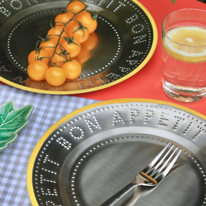 Two decorative steel plates with yellow trim embossed lettering and dotted borders, styled with food and a glass of lemon water on a red surface. One plate holds orange cherry tomatoes on the vine, and the other holds a fork.